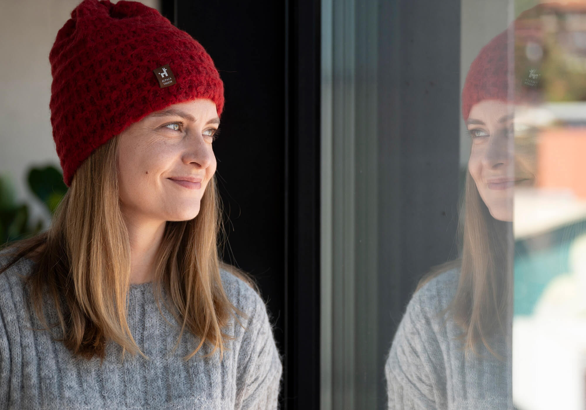 Frau blickt aus dem Fenster. Sie trägt einen weichen Alpaka Pullover und den Suri Loop Schal und Mütze aus seltener Suri Alpakawolle