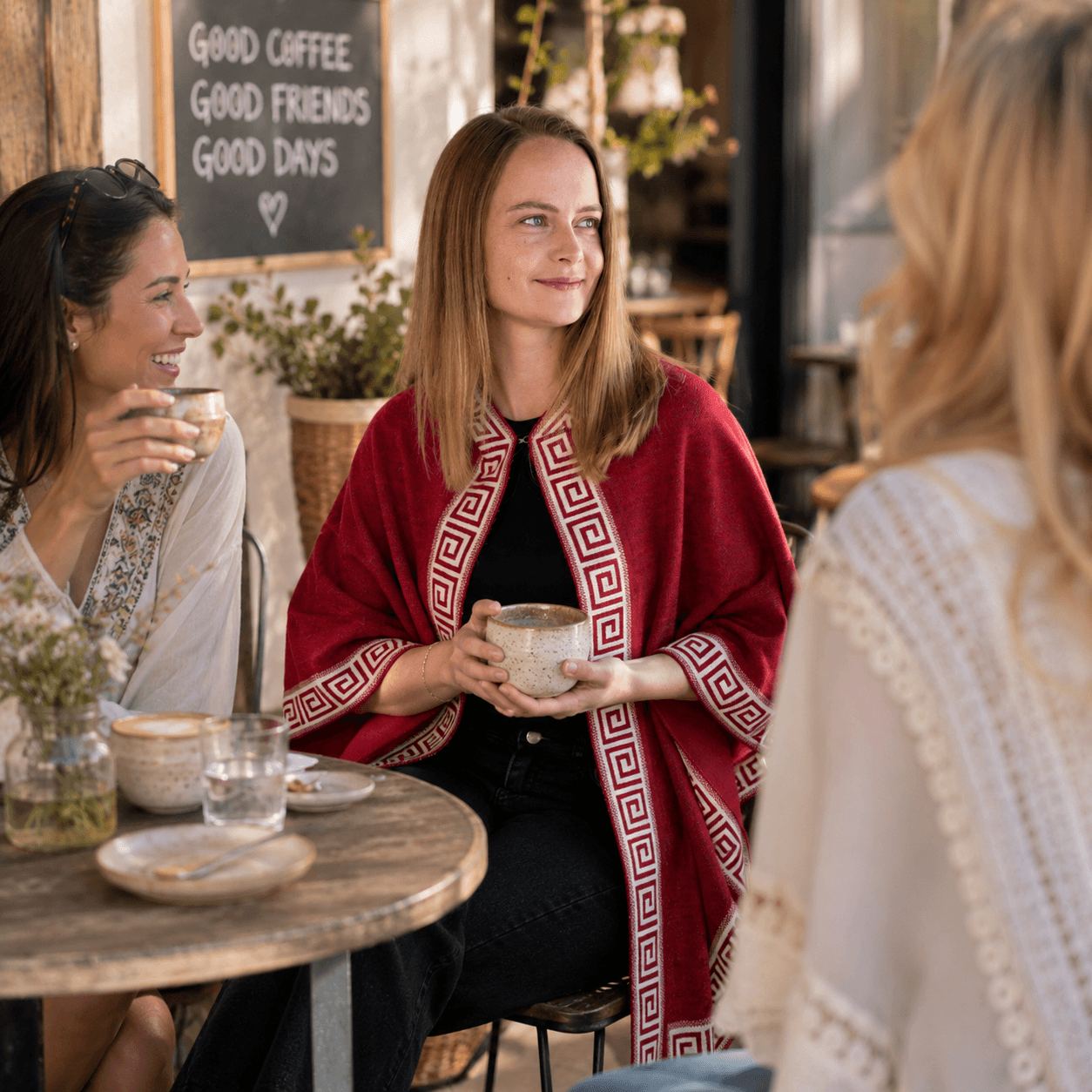 Frauen sitzen in einem Cafe, eine von ihnen trägt den Luz Alpaka Poncho mit eleganter Musterung in rot