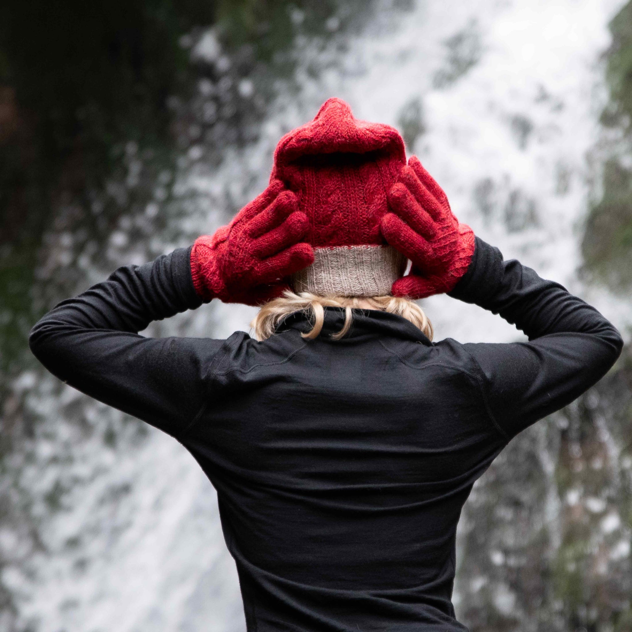 Model trägt vor Wasserfall Kulisse in Peru handgemachte Alpaka Wendehandschuhe für Damen und Herren von Alpakin in rot mit Mütze