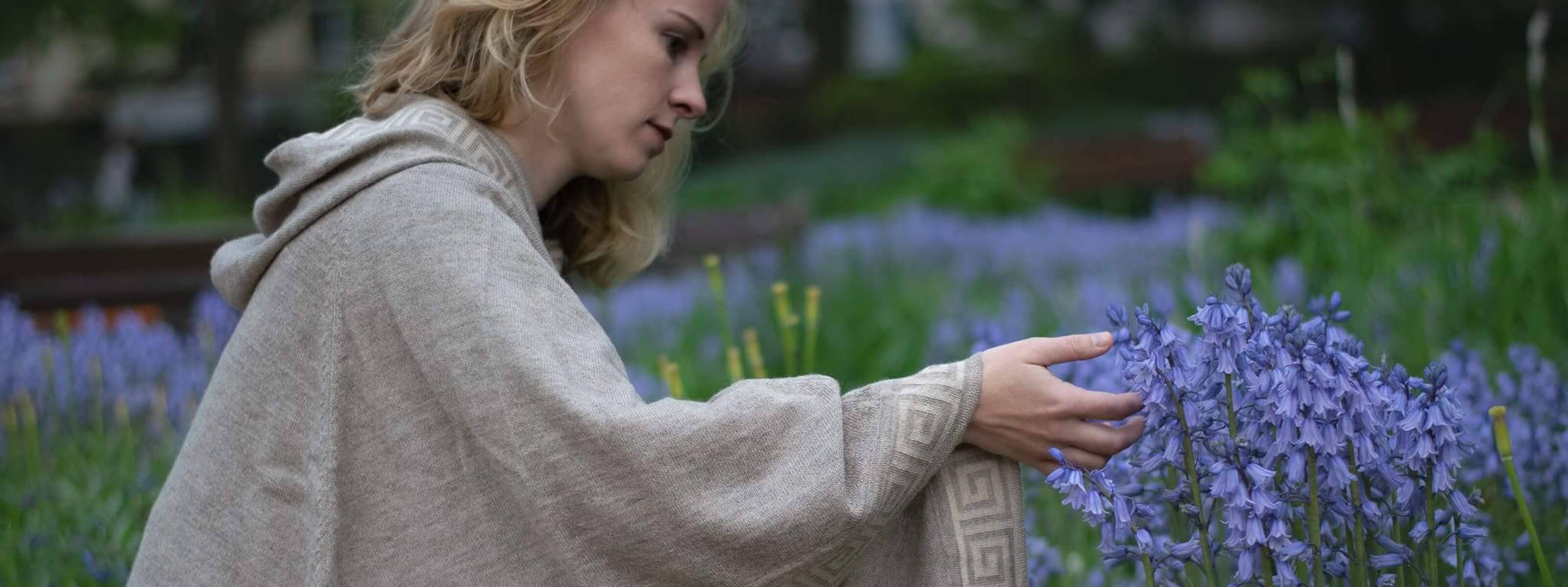 Frau vor Frühlingskullisse trägt den Luz Alpaka Poncho in beige und berührt Blumen