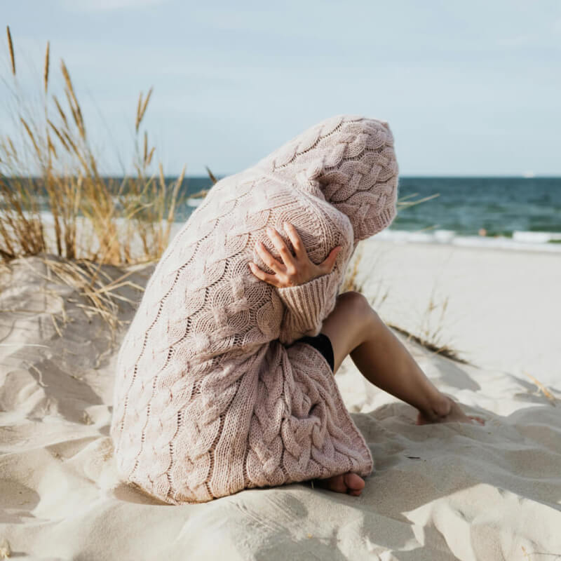 Frau sitzt am Strand und kuschelt sich in ihre dicke Strickjacke aus wärmster Alpakawolle